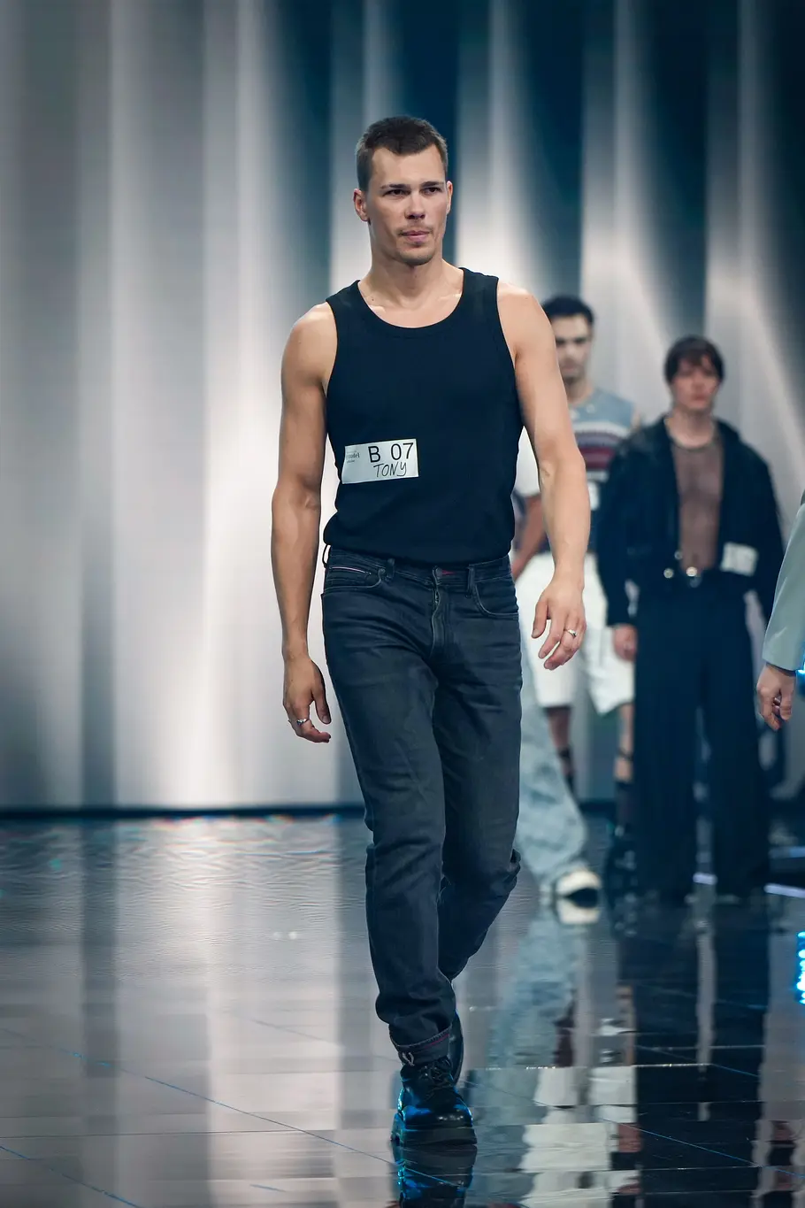 Ein männlicher Kandidat läuft bei „Germanys Next Topmodel“ selbstbewusst über den Catwalk. Hinter ihm stehen zahlreiche weitere männliche Kandidaten am Bühnenrand.
