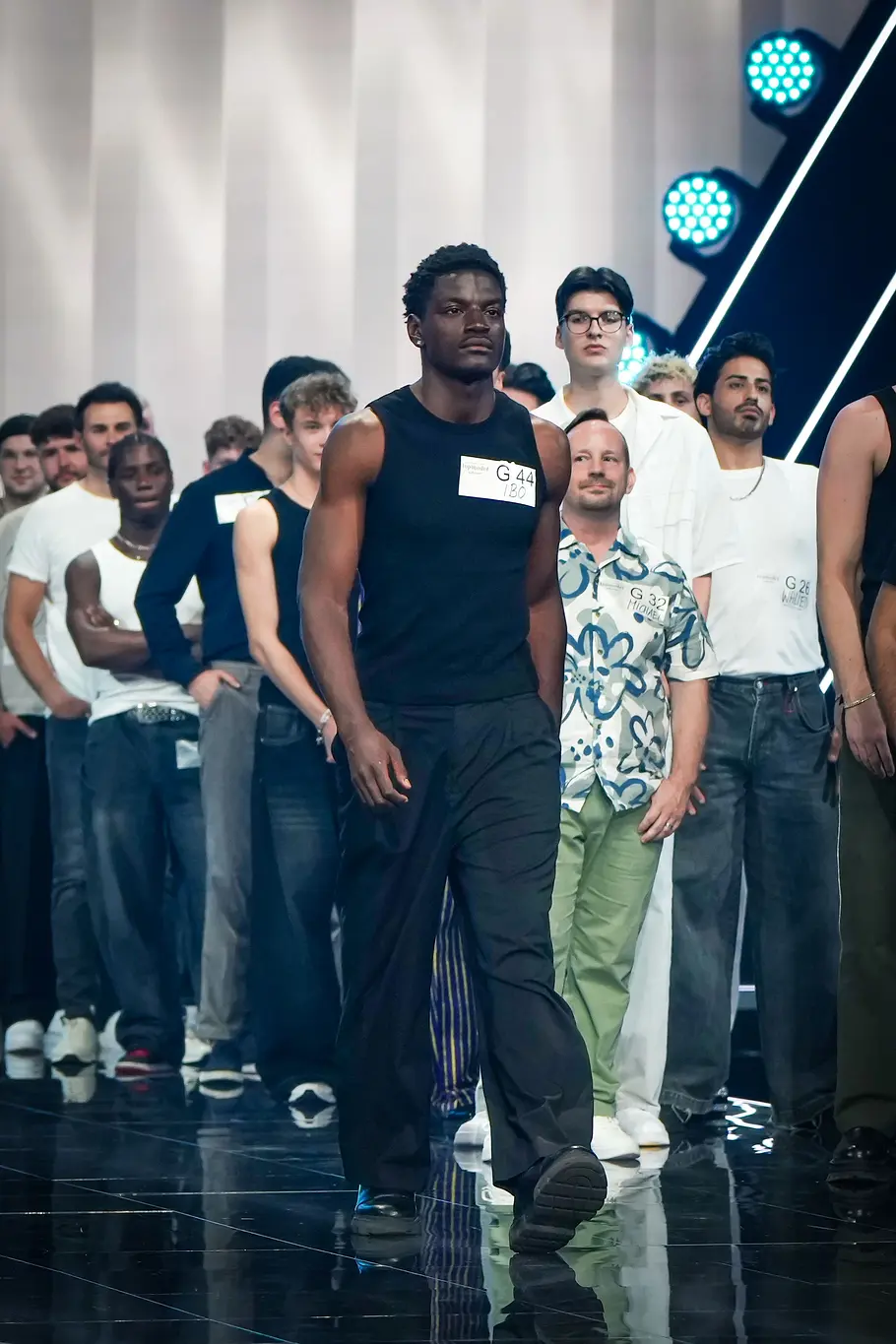 Ein männlicher Kandidat läuft bei „Germanys Next Topmodel“ selbstbewusst über den Catwalk. Hinter ihm stehen zahlreiche weitere männliche Kandidaten am Bühnenrand.
