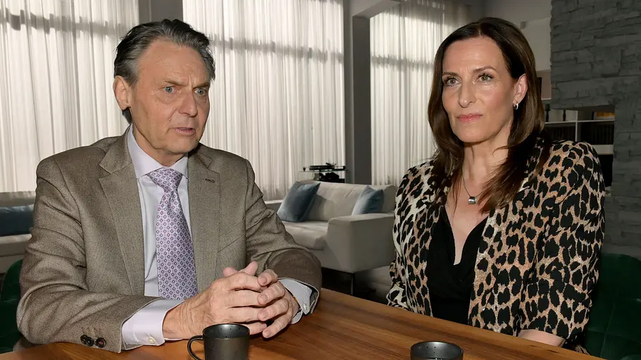 Kommt es zum Bruch zwischen Katrin und Gerner? Gerner im Anzug und Katrin mit Leopardenmuster-Jacke sitzen an einem Holztisch mit zwei schwarzen Tassen.