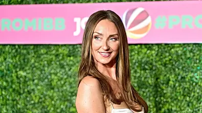 Gia Georgina lächelt bei der Verkündung der Promi Big Brother-Kandidaten 2026 in einem weiß-schwarz gemusterten Kleid vor einer grünen Pflanzenwand mit rosa Schriftbanner. - Foto: IMAGO / STAR-MEDIA