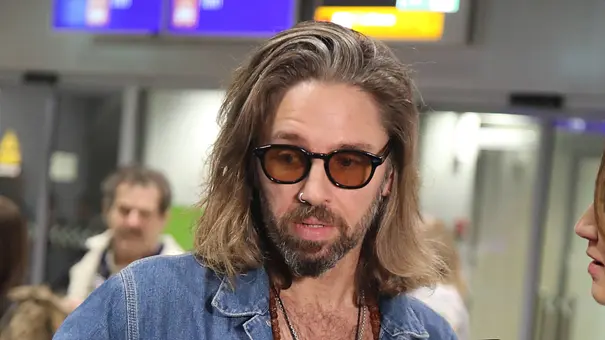 Langhaariger Mann mit Sonnenbrille und Jeanshemd gibt ein Interview vor TV-Mikrofonen in einem Flughafenbereich. - Foto: IMAGO / Matthias Wehnert