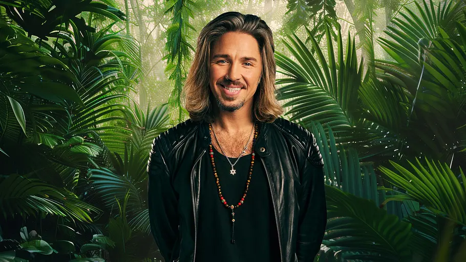 Gil Ofarim will im Dschungel 2026 sein Image verbessern. Gil Ofarim lächelt vor einem Dschungel-Hintergrund.