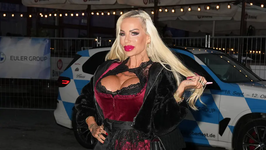 Gina-Lisa auf dem Oktoberfest 2025 Gina-Lisa in rotem Dirndl und schwarzem Mantel posiert vor einem geparkten Fahrzeug bei Nacht.