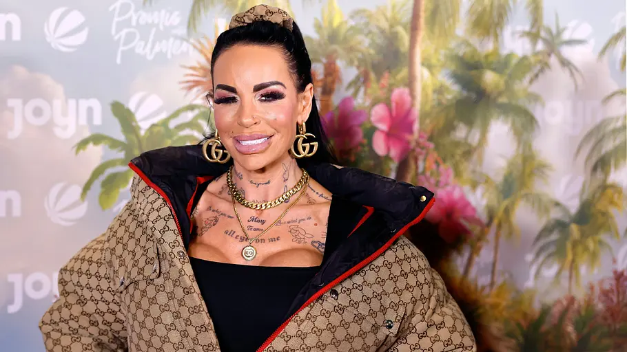 Gina-Lisa bei der Pup-Premiere mit Hochsteckfrisur, auffälligem Schmuck und gemusterter Jacke lächelt vor einer tropischen Fotowand mit Logos.