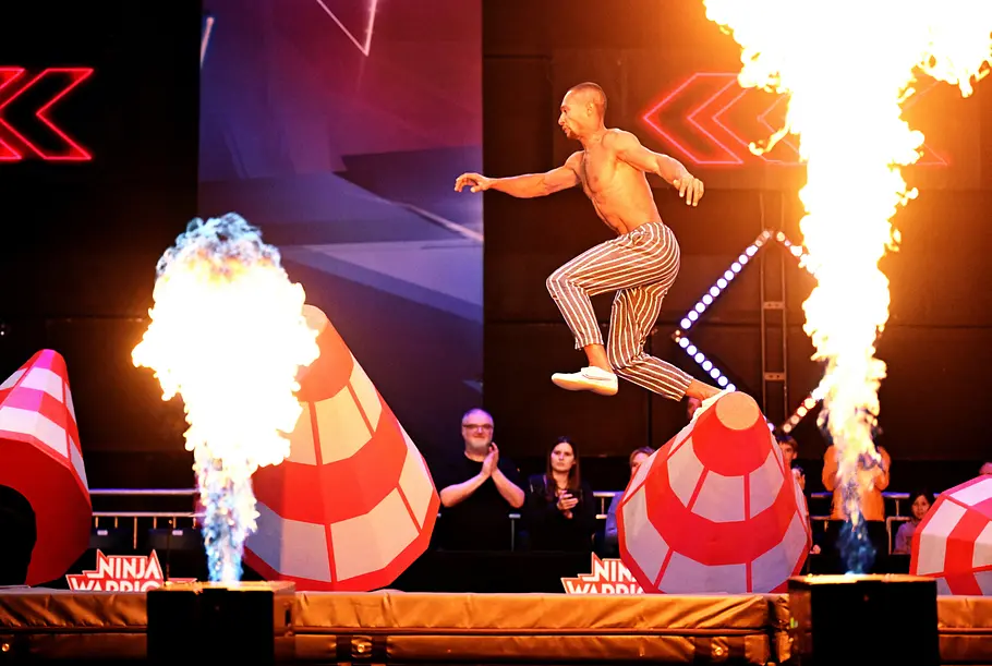 Giovanni Ertl ist Teilnehmer bei Ninja Warrior Germany 2026 Giovanni Ertl springt mit freiem Oberkörper und gestreifter Hose zwischen roten Hindernissen. Daneben lodern hohe Flammen.