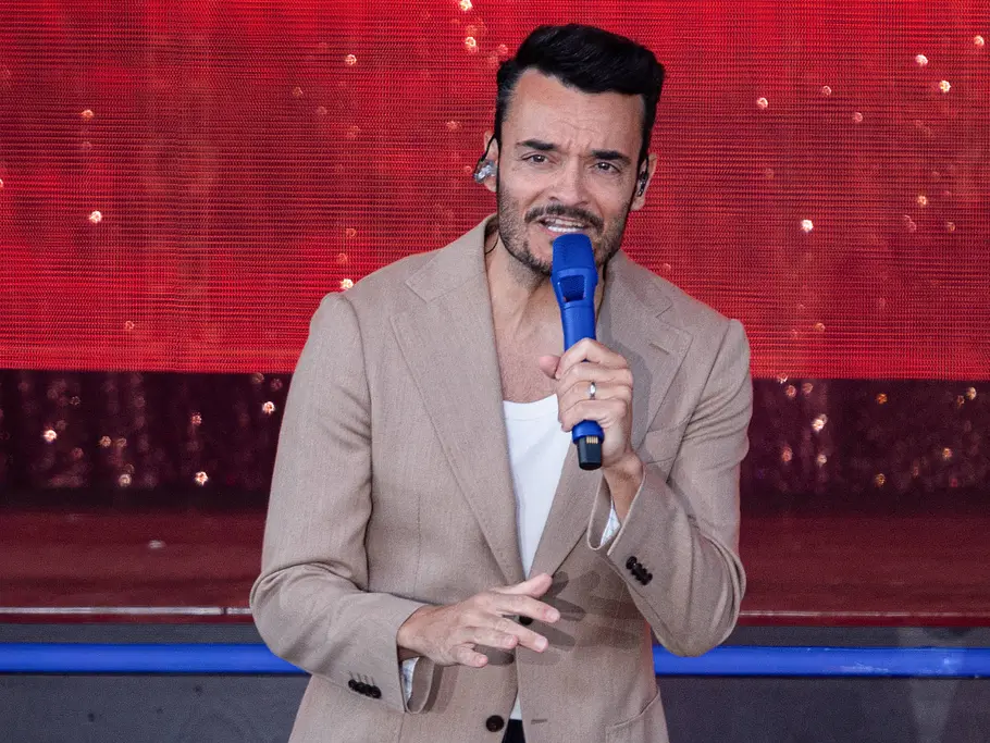 Giovanni Zarrella bei der „Helene Fischer Show“ 2026 Giovanni Zarrella steht im beigen Sakko mit einem blauen Mikrofon vor einem rot beleuchteten Bühnenhintergrund.