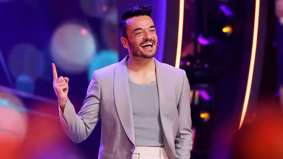Giovanni Zarrella Die große Weihnachtsshow Giovanni Zarrella lacht auf einer bunt beleuchteten Bühne und hebt einen Zeigefinger. Er trägt ein helles Sakko.