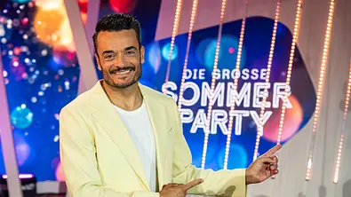 Die Giovanni Zarrella Show - Die große Sommerparty: Moderator Giovanni Zarrella lächelt in die Kamera und zeigt mit beiden Zeigefingern auf das Sendungslogo Die große Sommerparty. Er trägt einen cremefarbenen Anzug und ein weißes T-Shirt. - Foto: ZDF/Sascha Baumann Die Giovanni Zarrella Show - Die große Sommerparty: Moderator Giovanni Zarrella lächelt in die Kamera und zeigt mit beiden Zeigefingern auf das Sendungslogo Die große Sommerparty. Er trägt einen cremefarbenen Anzug und ein weißes T-Shirt. - Foto: ZDF/Sascha Baumann