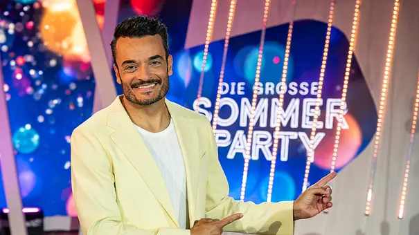 Die Giovanni Zarrella Show - Die große Sommerparty: Moderator Giovanni Zarrella lächelt in die Kamera und zeigt mit beiden Zeigefingern auf das Sendungslogo Die große Sommerparty. Er trägt einen cremefarbenen Anzug und ein weißes T-Shirt. - Foto: ZDF/Sascha Baumann Die Giovanni Zarrella Show - Die große Sommerparty: Moderator Giovanni Zarrella lächelt in die Kamera und zeigt mit beiden Zeigefingern auf das Sendungslogo Die große Sommerparty. Er trägt einen cremefarbenen Anzug und ein weißes T-Shirt. - Foto: ZDF/Sascha Baumann