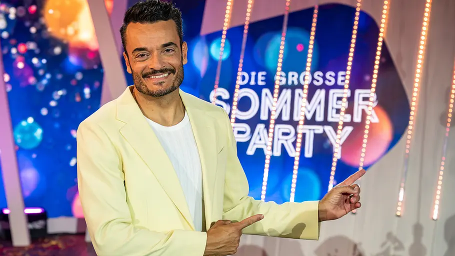 „Giovanni Zarrella Show“: Hinter den Kulissen wird es plötzlich hektisch Die Giovanni Zarrella Show - Die große Sommerparty: Moderator Giovanni Zarrella lächelt in die Kamera und zeigt mit beiden Zeigefingern auf das Sendungslogo Die große Sommerparty. Er trägt einen cremefarbenen Anzug und ein weißes T-Shirt.