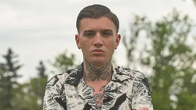 Ein junger Mann mit kurzen, zurückgekämmten Haaren und zahlreichen Tattoos an Hals und Armen steht mit verschränkten Armen auf einer grünen Wiese. Er trägt ein kurzärmliges, schwarz-weiß gemustertes Hemd und dunkle Hosen. Im Hintergrund sind Bäume und ein unscharfer Naturbereich zu sehen. - Foto: RTL