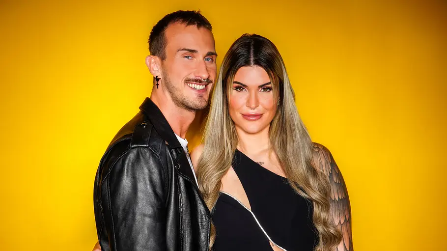 Gloria Schwesinger und Michael Gloria Schwesinger im schwarzen Kleid und Michael in schwarzer Lederjacke stehen eng beieinander vor gelbem Hintergrund.