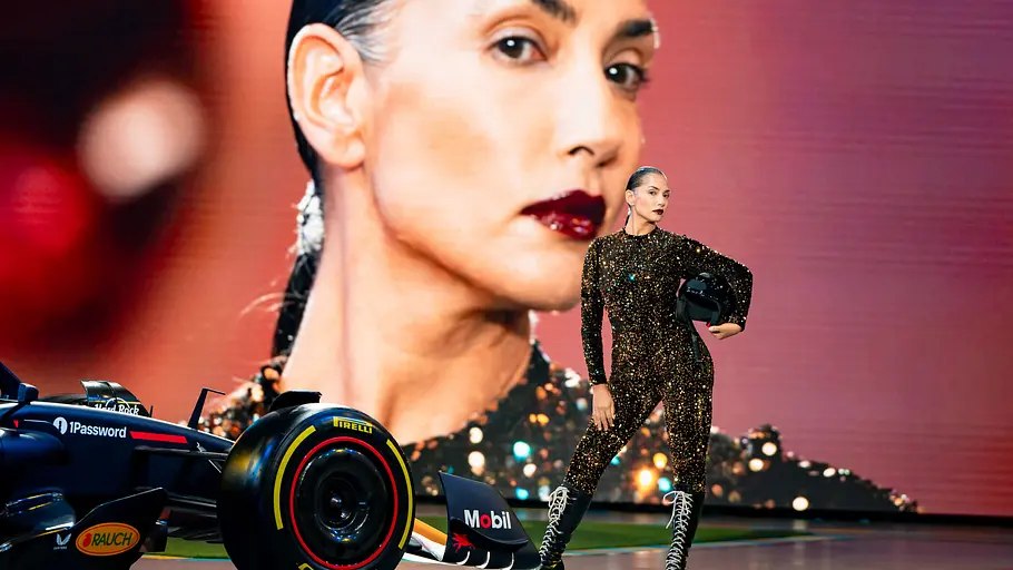 Bianca beim Entscheidungswalk Model in einem gold-schwarz funkelnden Jumpsuit und hohen Schnürstiefeln steht mit einem Helm in der Hand neben einem Formel-1-Rennwagen, im Hintergrund eine große Porträtprojektion ihres Gesichts.