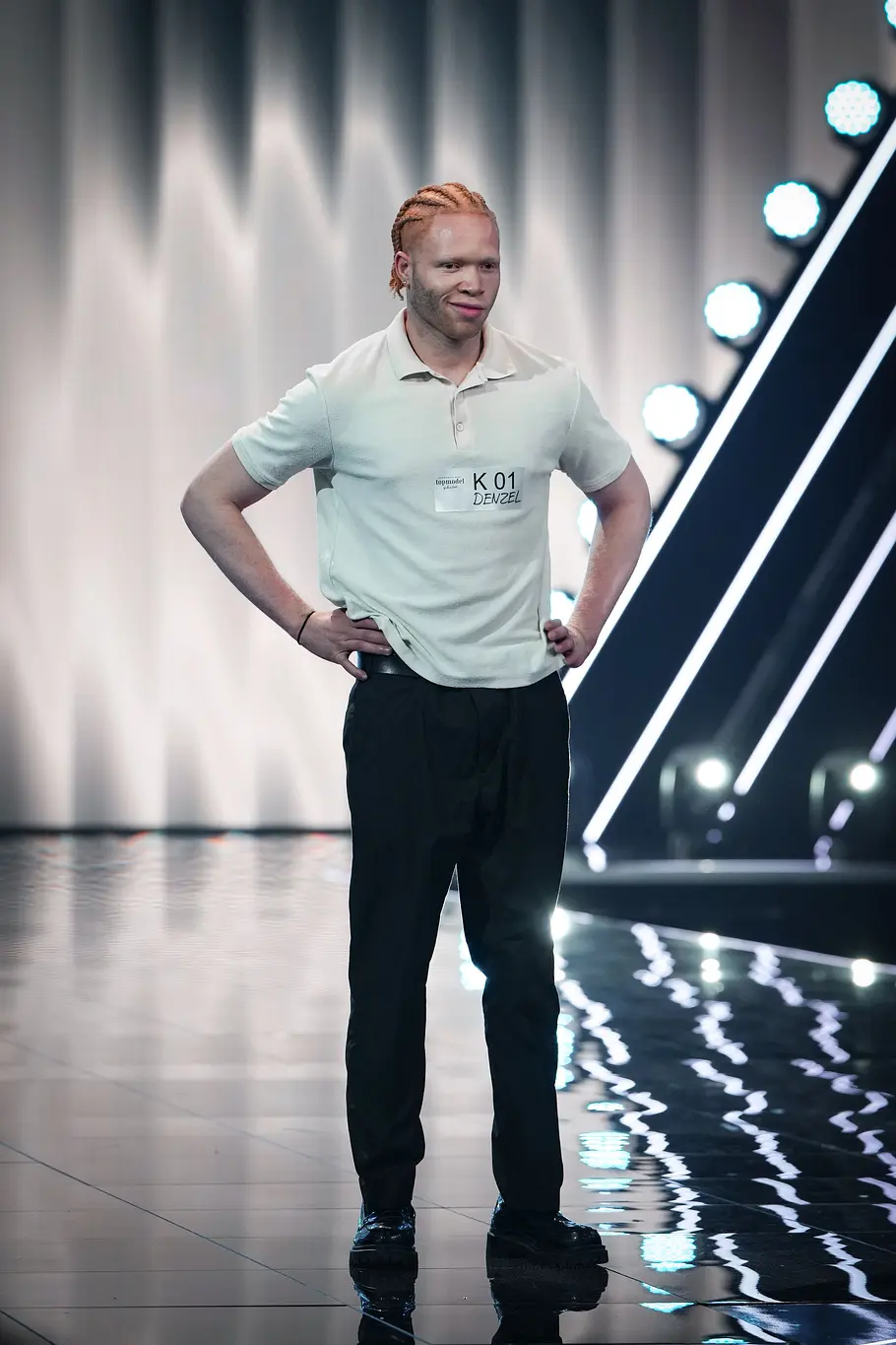 Ein Kandidat mit geflochtenen, orangefarbenen Haaren steht mit den Händen in die Hüften gestützt auf dem Laufsteg und trägt ein helles Poloshirt sowie schwarze Hose.