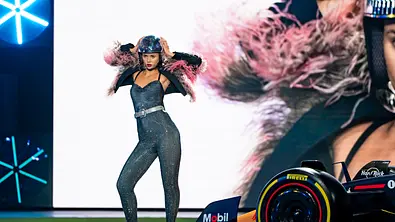 Model in einem glitzernden, eng anliegenden Jumpsuit mit Federdetails an den Schultern und Helm posiert selbstbewusst auf einem Laufsteg neben einem Formel-1-Rennwagen. - Foto: ProSieben/Daniel Graf
