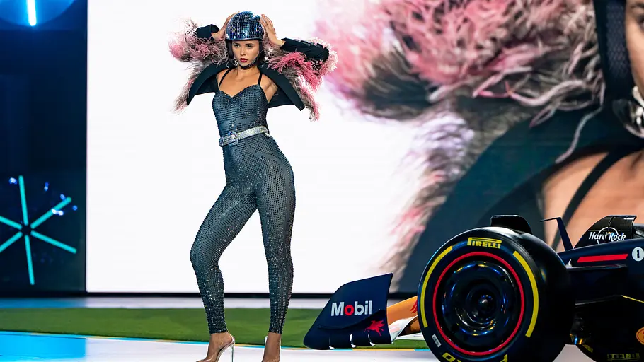 GNTM Entscheidungswalk Model in einem glitzernden, eng anliegenden Jumpsuit mit Federdetails an den Schultern und Helm posiert selbstbewusst auf einem Laufsteg neben einem Formel-1-Rennwagen.