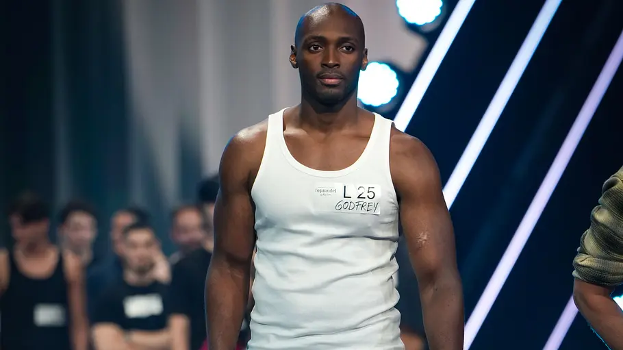 Godfrey beim GNTM Casting Ein großer, muskulöser Mann steht auf einem Catwalk. Er trägt ein weißes Tanktop mit dem Namensschild L 25 GODFREY und eine blaue Jeans. Im Hintergrund stehen weitere Personen, leicht unscharf und im Schatten.