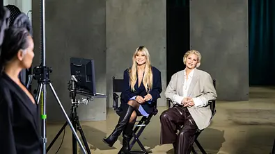 Zwei Frauen sitzen in einem Studio auf Regiestühlen vor einer Kamera und einem Monitor, während im Vordergrund eine weitere Person von der Seite zu sehen ist. - Foto: ProSieben/Daniel Graf