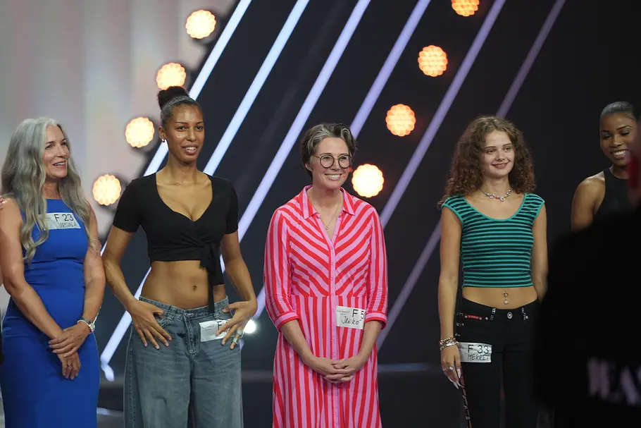Ildikó von Kürthy wagt das Casting Eine Gruppe von fünf weiblichen Teilnehmerinnen steht nebeneinander auf einem Catwalk und lächelt. Die Frauen tragen verschiedene Outfits von modern bis elegant, darunter ein blaues Kleid, ein schwarzes Oberteil mit weiten Jeans, ein pink-weiß gestreiftes Kleid, ein grün-schwarz gestreiftes Top und eine schwarze Hose. Im Hintergrund sind Lichter und ein moderner Bühnenaufbau zu sehen.
