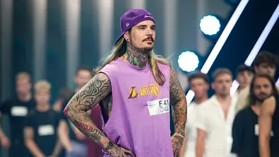 GNTM-Kandidat Jill steht beim Entscheidungswalk auf dem Laufsteg und trägt ein lilafarbenes Outfit mit Cap, während andere Models im Hintergrund warten. - Foto:  ProSieben/Daniel Graf