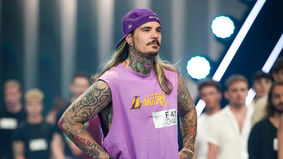 Jill bei GNTM GNTM-Kandidat Jill steht beim Entscheidungswalk auf dem Laufsteg und trägt ein lilafarbenes Outfit mit Cap, während andere Models im Hintergrund warten.