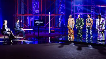 Bühnenbild von „Germany’s Next Topmodel“ mit Jury am Tisch und vier männlichen Models auf dem Laufsteg unter blau-roter Studiobeleuchtung. - Foto: ProSieben/Daniel Graf