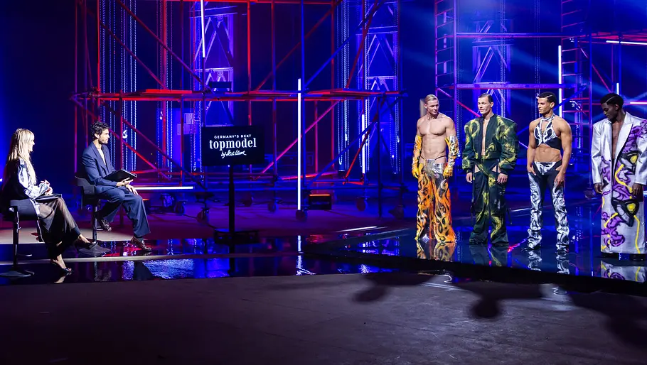 GNTM-Model verschwunden? Bühnenbild von „Germany’s Next Topmodel“ mit Jury am Tisch und vier männlichen Models auf dem Laufsteg unter blau-roter Studiobeleuchtung.