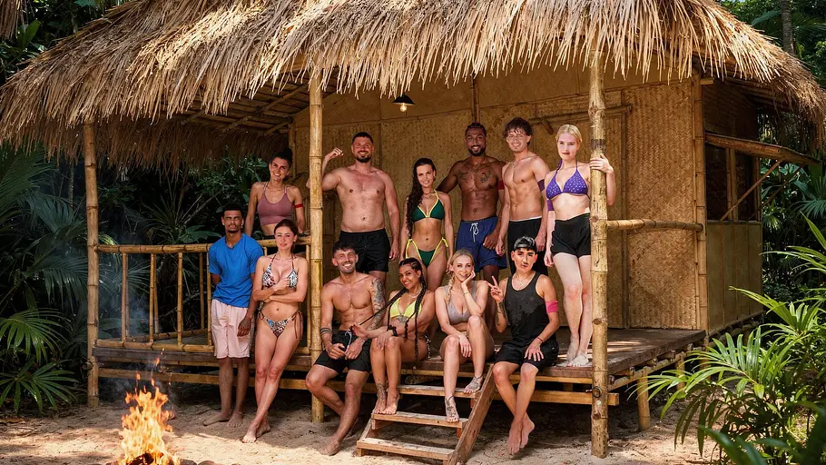 Good Luck Guys Die Teilnehmer:innen der Realityshow „Good Luck Guys“ posieren gemeinsam vor einer strohgedeckten Dschungelhütte. Die Gruppe trägt überwiegend Badebekleidung und Sommerkleidung, einige sitzen auf den Stufen der Hütte, andere stehen auf der Veranda. Im Vordergrund brennt ein Lagerfeuer, während dichter tropischer Wald die Kulisse umgibt.