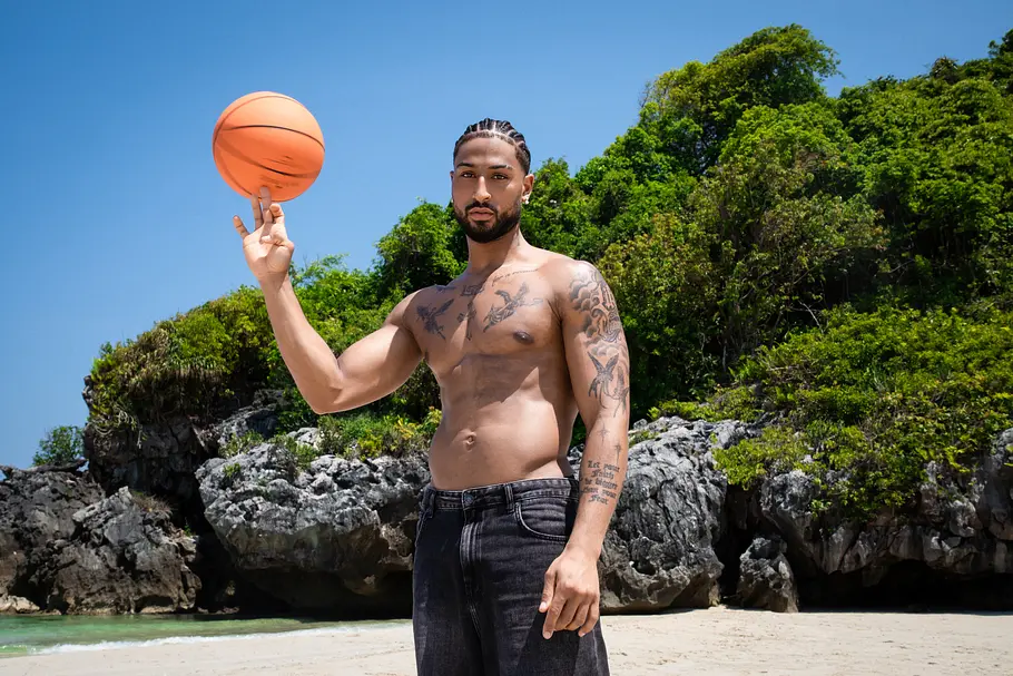 Calvin Ogara Reality-TV-Teilnehmer Calvin Ogara steht oberkörperfrei an einem tropischen Strand und balanciert einen Basketball auf seinem Finger. Im Hintergrund sind Felsen, dichter grüner Dschungel sowie der helle Sandstrand unter blauem Himmel zu sehen.