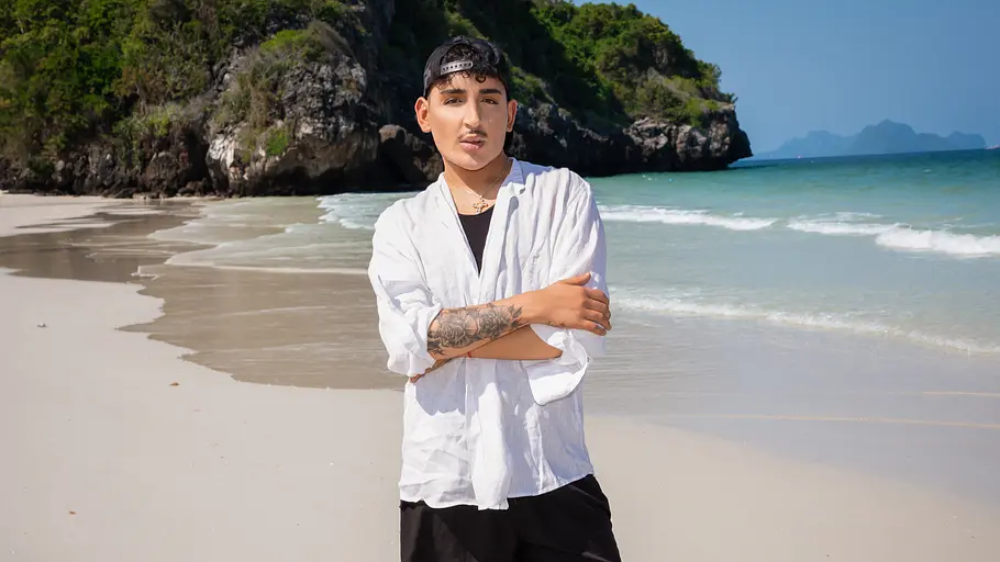 Francesco Morelli Francesco Morelli posiert mit verschränkten Armen in weißem Hemd und schwarzer Shorts an einem tropischen Sandstrand vor türkisblauem Meer bei „Good Luck Guys“.