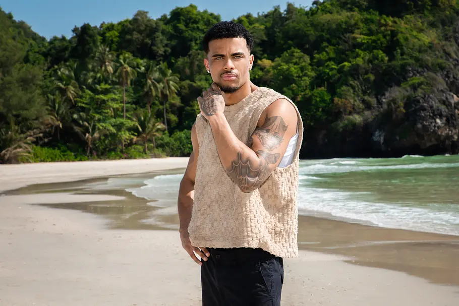 Dion Brown Reality-TV-Teilnehmer Dion Brown posiert an einem tropischen Sandstrand vor dichter Dschungelkulisse. Er trägt ein ärmelloses, beigefarbenes Oberteil und dunkle Shorts, während seine tätowierten Arme gut sichtbar sind. Im Hintergrund sind Palmen, das türkisfarbene Meer und sanfte Wellen zu sehen.