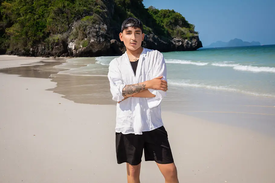 Zoello Boy Reality-TV-Teilnehmer Francesco Morelli steht mit verschränkten Armen an einem tropischen Sandstrand. Er trägt ein weißes Leinenhemd, schwarze Shorts und eine rückwärts getragene Kappe. Im Hintergrund sind türkisblaues Meer, Felsen und dichter Dschungel zu sehen.