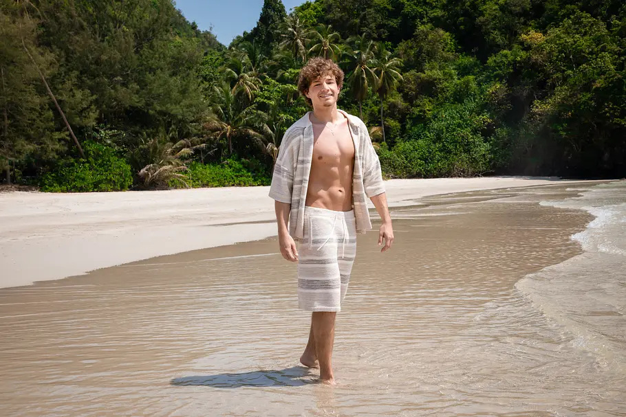 Garry Secret Reality-TV-Teilnehmer Garry Secret läuft barfuß durch das flache Wasser an einem tropischen Sandstrand. Er trägt ein helles, offenes Sommerhemd mit passender Shorts und lächelt in die Kamera. Im Hintergrund sind Palmen, dichter Dschungel und die ruhige Meeresküste zu sehen.