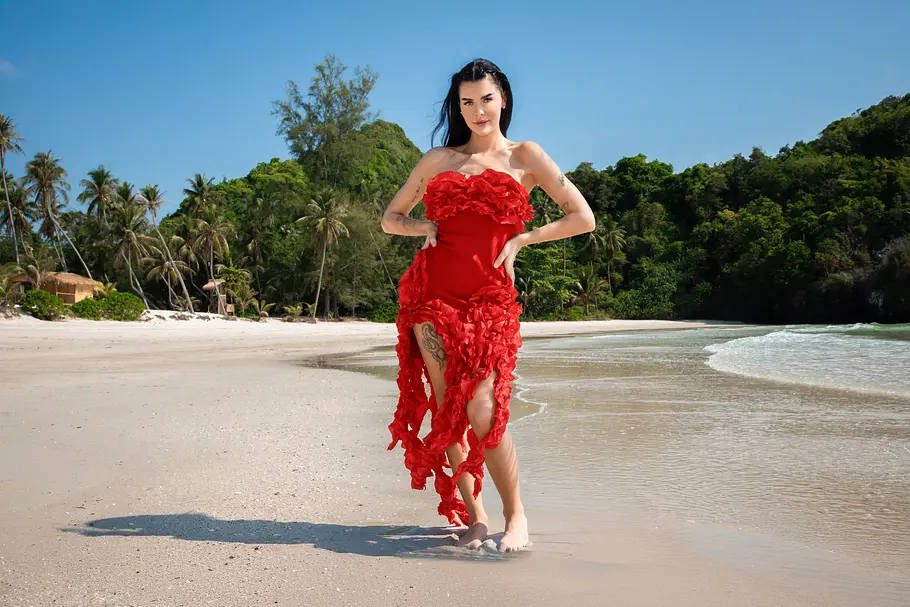 Nelly Reality-TV-Teilnehmerin Nelly läuft barfuß an einem tropischen Sandstrand entlang. Sie trägt ein auffälliges rotes Kleid mit floralen Applikationen und blickt selbstbewusst in die Kamera. Im Hintergrund sind Palmen, dichter Dschungel, heller Sand und das türkisfarbene Meer zu sehen.