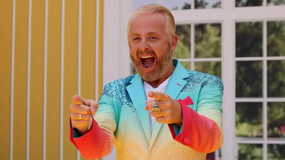 Ross Antony zeigt in einem bunten Anzug lachend in die Ferne. 
