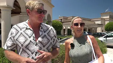 Steff und Peggy stehen vor einer luxuriösen Hotel- oder Ferienanlage im Freien. Beide tragen Sonnenbrillen und sommerliche Kleidung, während sie sich unterhalten. Im Hintergrund sind Palmen, gepflegte Grünanlagen und mehrere Gebäude zu sehen. - Foto: RTL
