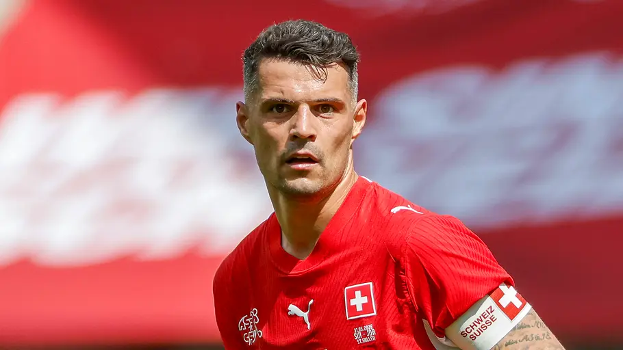 Ein Fußballspieler der Schweizer Nationalmannschaft im roten Trikot mit der Rückennummer 10 und Kapitänsbinde blickt während eines Spiels konzentriert zur Seite. Im Hintergrund ist unscharf eine große Schweizer Flagge zu sehen.