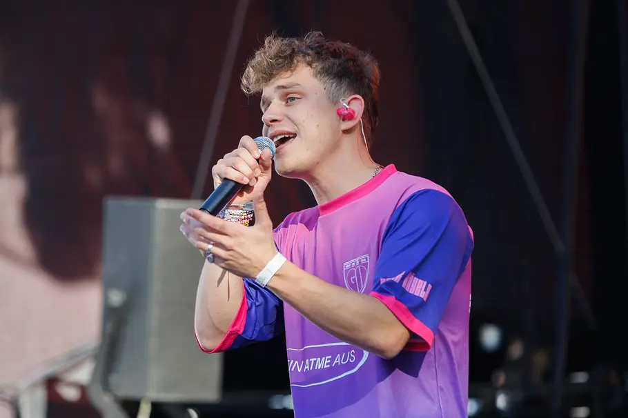 Gregor Hägele ZDF Weihnachtsshow Gregor Hägele singt auf einer Bühne in ein Handmikrofon. Er trägt ein lila-blaues Shirt und pinke In-Ear-Kopfhörer.