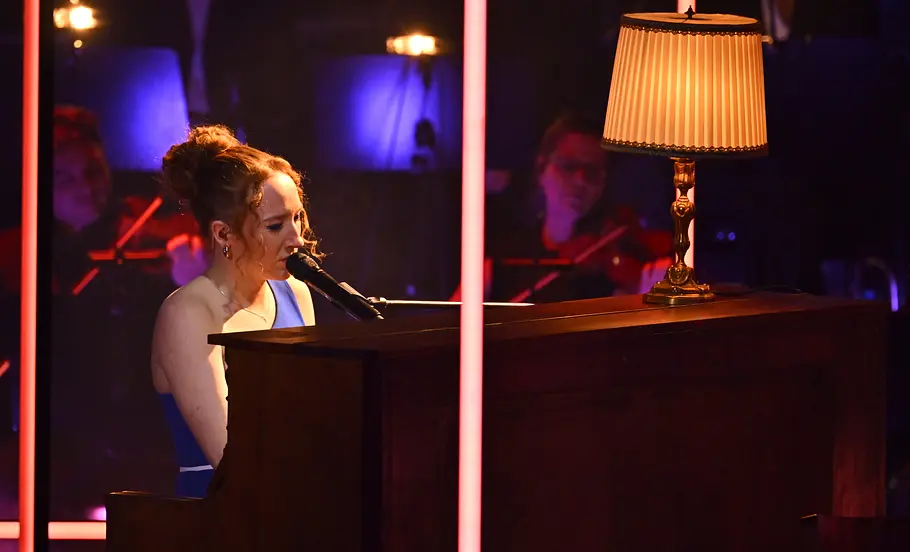 Greta Heimann Halbfinale The Voice of Germany 2025 Greta Heimann am Klavier. Ein gemütliche Lampe steht auf dem Klavier. Sie trägt ein blaues Kleid.