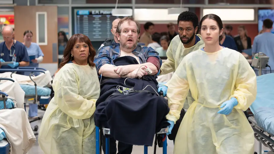 Greys Anatomy: Ausnahmezustand im Grey Sloan Memorial Drei Personen in gelben Schutzkitteln begleiten einen Patienten mit Sauerstoffschlauch auf einer Liege durch eine Notaufnahme.