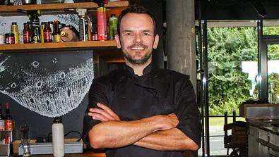 Szeffen Henssler steht in einer Küche und verschränkt breit lächelnd die Arme vor seinem Körper. - Foto: IMAGO / BREUEL-BILD