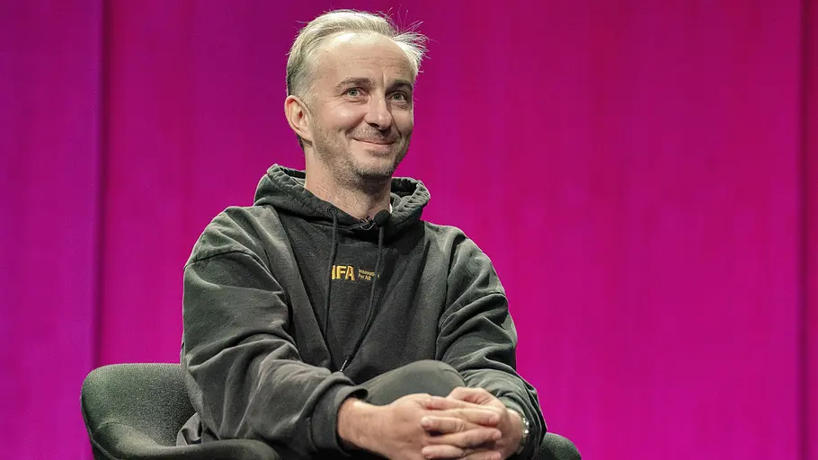 Gründe Absetzung „ZDF Magazin Royale“ Jan Böhmermann sitzt lächelnd im dunklen Kapuzenpullover mit verschränkten Händen in einem Sessel vor pinkem Hintergrund.