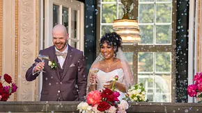 Gunnar und Josy stehen lächelnd in Hochzeitskleidung mit Sektgläsern auf einem blumengeschmückten Balkon voller Seifenblasen. - Foto: © Netflix Gunnar und Josy stehen lächelnd in Hochzeitskleidung mit Sektgläsern auf einem blumengeschmückten Balkon voller Seifenblasen. - Foto: © Netflix
