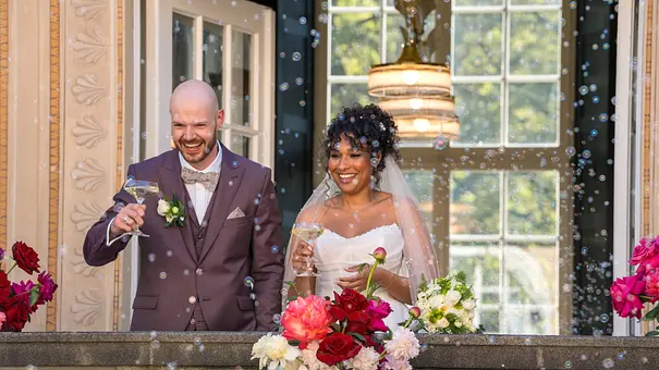 Gunnar und Josy stehen lächelnd in Hochzeitskleidung mit Sektgläsern auf einem blumengeschmückten Balkon voller Seifenblasen. - Foto: © Netflix Gunnar und Josy stehen lächelnd in Hochzeitskleidung mit Sektgläsern auf einem blumengeschmückten Balkon voller Seifenblasen. - Foto: © Netflix