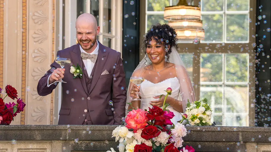 Gunnar und Josy sind ein Beispiel dafür, dass es bei „Love is Blind“ echte Happy Ends geben kann. Gunnar und Josy stehen lächelnd in Hochzeitskleidung mit Sektgläsern auf einem blumengeschmückten Balkon voller Seifenblasen.