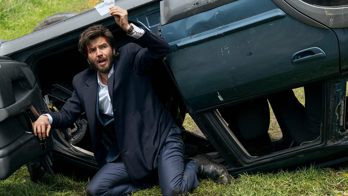 Guy Burnet als Theodore Chips in „Fubar“ Staffel 2. Theodore Chips kniet vor der geöffneten Tür eines Autos, das auf dem Dach liegt. Er hebt mit der linken Hand einen Zettel über seinen Kopf.