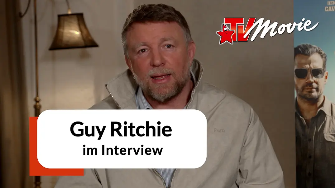 Guy Ritchie sitzt für ein Interview in einem Hotelzimmer. Hinter ihm ist ein Plakat des Films „In the Grey“.  - Foto: Leonine/TVMovie