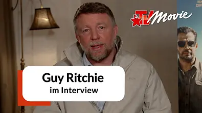 Guy Ritchie sitzt für ein Interview in einem Hotelzimmer. Hinter ihm ist ein Plakat des Films „In the Grey“.  - Foto: Leonine/TVMovie