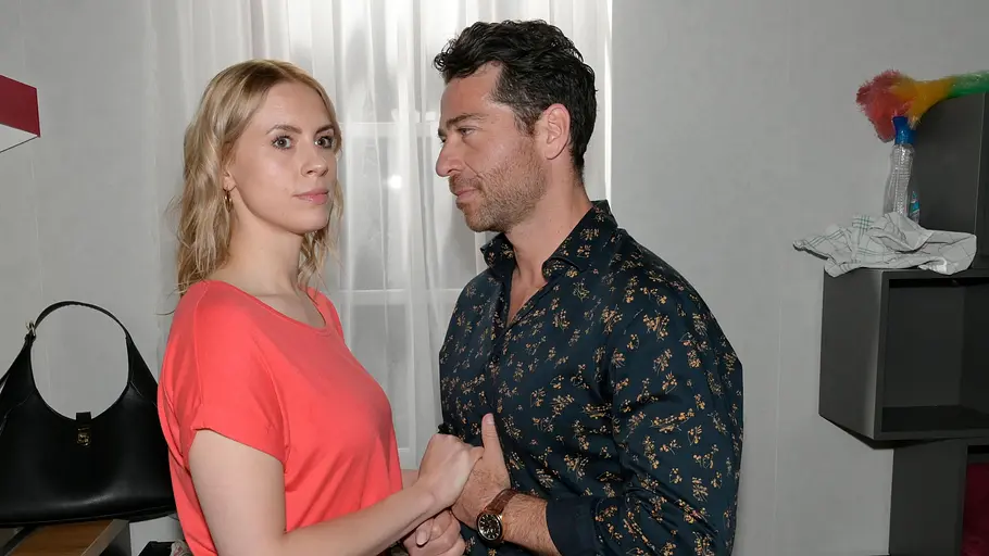 Zoe erfährt Neues über Carlos. Carlos schaut Zoe an und hält ihre Hand, während sie ernst zur Seite blickt.
