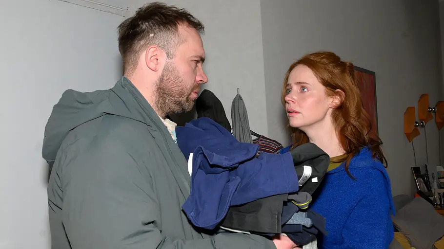 Toni schmeißt Erik raus. Toni schaut Erik wütend an, in der Hand hält sie seine Sachen.